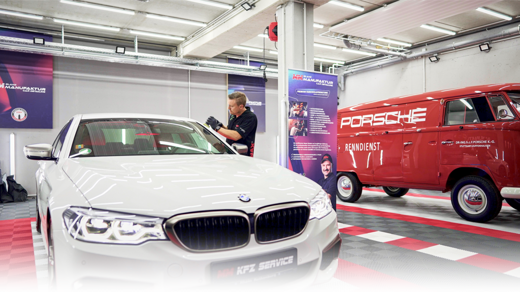 Das Bild zeigt Manuel Muhr in seiner Werkstatt. Er poliert einen weißen BMW. Im Hintergrund steht ein roter VW Bully mit der Aufschrift "Porsche Renndienst"