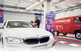 Das Bild zeigt Manuel Muhr in seiner Werkstatt. Er poliert einen weißen BMW. Im Hintergrund steht ein roter VW Bully mit der Aufschrift "Porsche Renndienst"