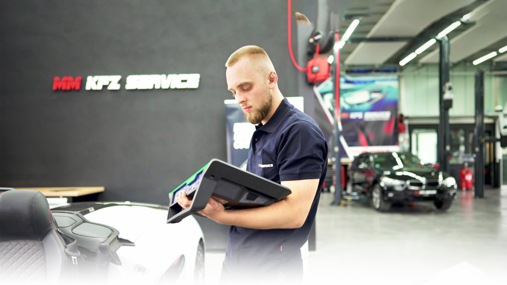 Das Foto zeigt einen KFZ-Mechaniker bei MM KFZ Service in der Werkstatt. Er arbeitet an einem weißen Audi R8. Im Hintergrund befindet sich ein schwarzer BMW.