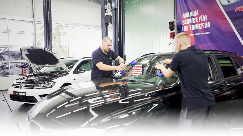 Das Bild zeigt zwei KFZ Mechaniker, die in der MM KFZ Werkstatt eine Glasscheibe an einem Auto tauschen